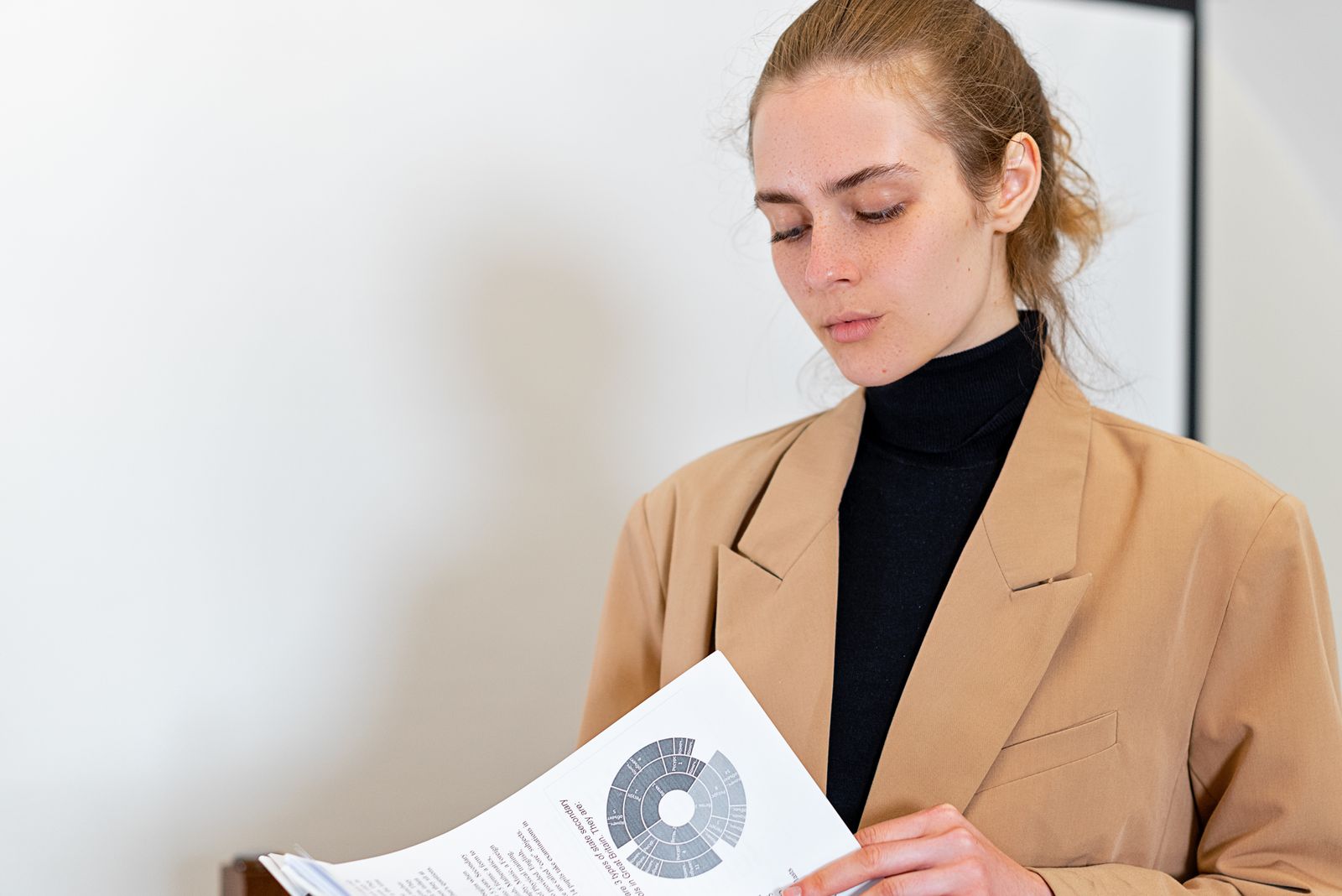 Eine Frau in camelfarbenem Blazer und schwarzem Rollkragenpullover blickt auf einen ausgedruckten Bericht mit einem Diagramm.