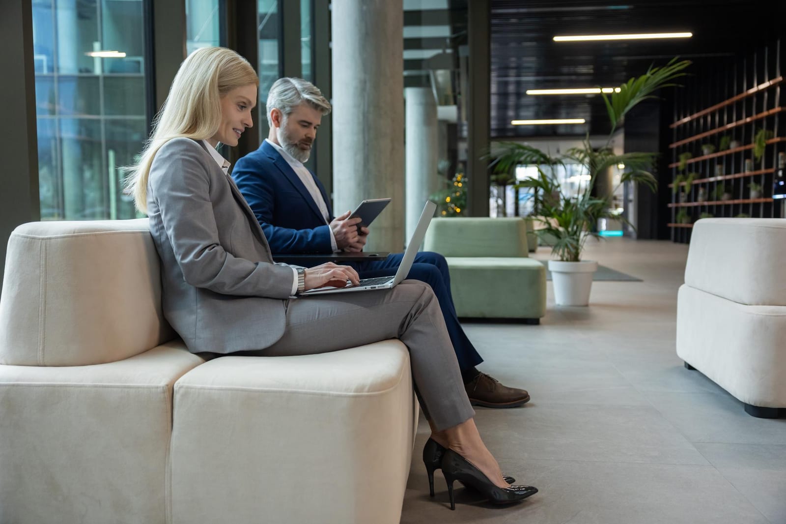 Eine Frau und ein Mann in Businesskleidung sitzen auf einem Sofa in einer Lobby, sie am Laptop, er am Tablet.